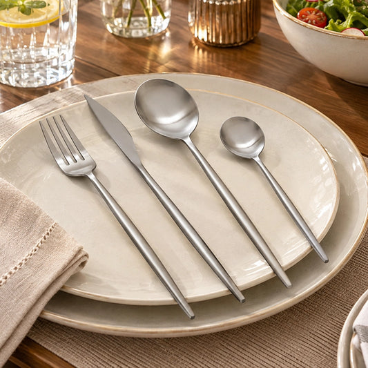 16 piece silver stainless steel cutlery set arranged on a white dinner plate with a wooden dining table backdrop