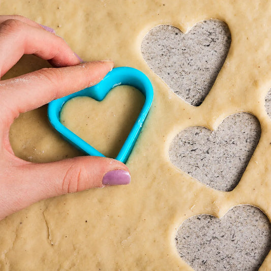 Hand holding a blue heart shape cookie cutter above rolled dough with cut heart cookies