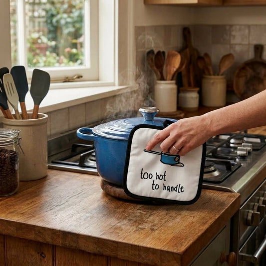 Blue cotton pot holder printed with "too hot to handle" gripping a cast iron pot on a gas hob in a bright kitchen