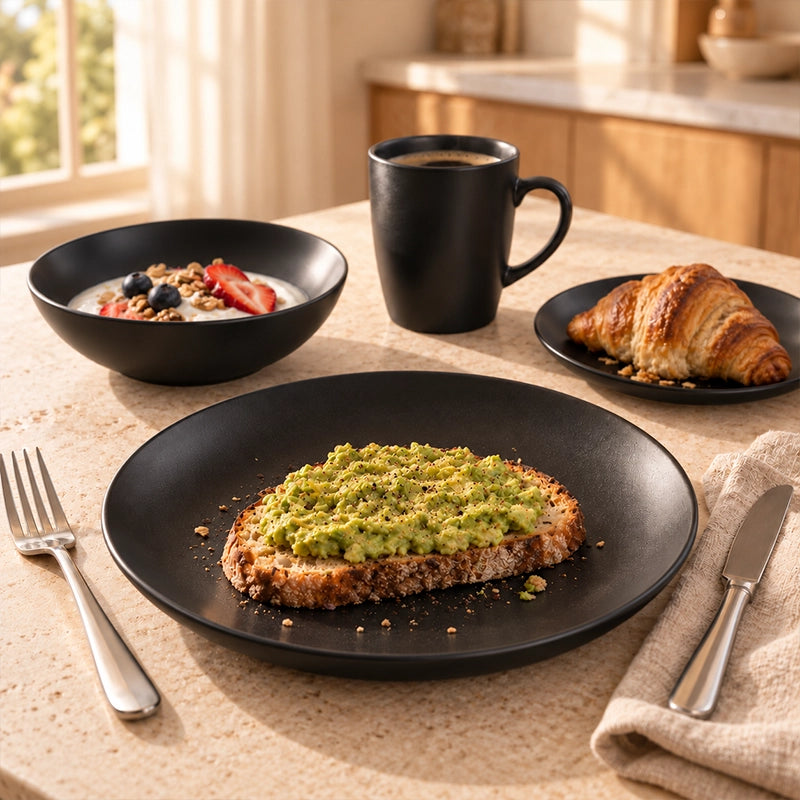 Matte black 12-piece stoneware dinner set with plate, bowl and mug served with avocado toast and a croissant on a linen table