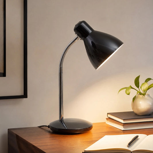 Black desk lamp with warm light on wooden table beside books and plant