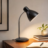 Black desk lamp with warm light on wooden table beside books and plant