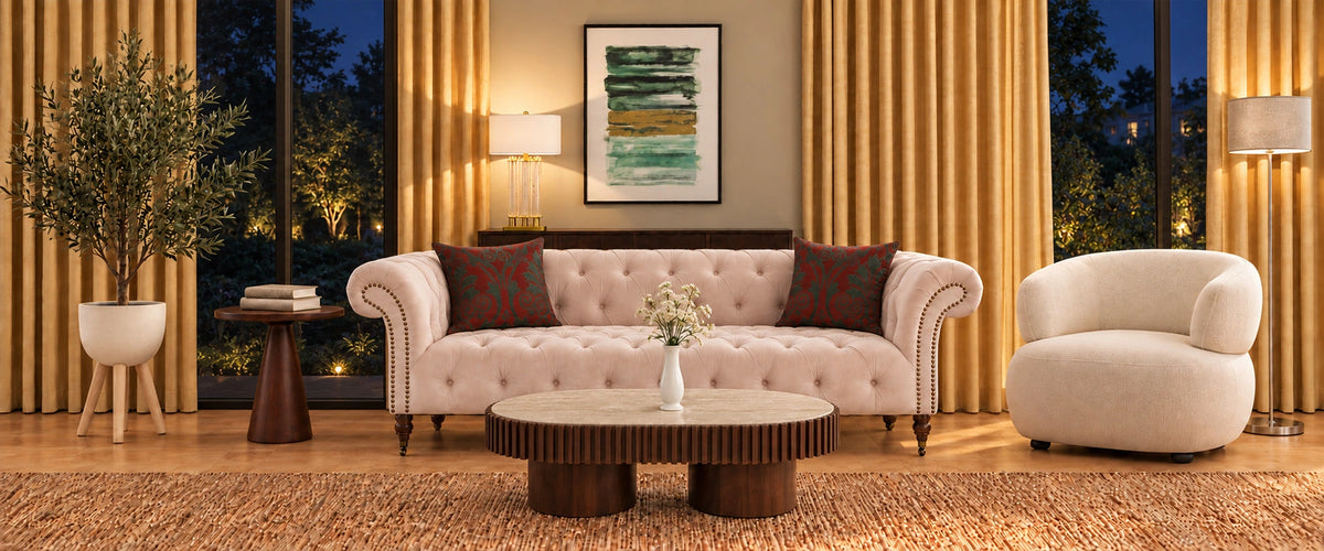 A styled living room scene featuring a tufted blush pink sofa with dark red patterned pillows, a round ribbed coffee table, a cream armchair, gold curtains, and a warm wood floor.