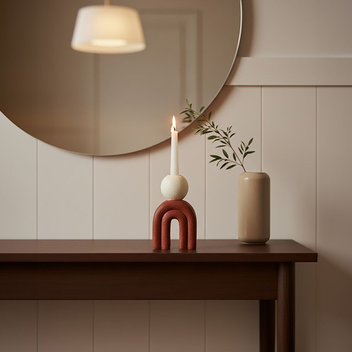 Red sculptural candle holder with white taper candle displayed beneath a round wall mirror