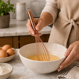 Rose gold kitchen whisk mixing batter in a ceramic bowl on a kitchen counter