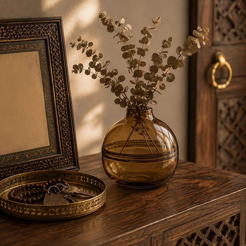 Small brown round glass vase with eucalyptus stems styled on a carved wooden console table