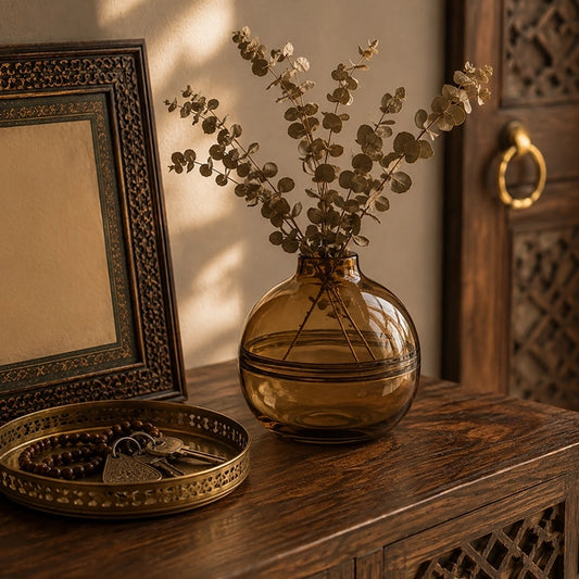 Small brown round glass vase with eucalyptus stems styled on a carved wooden console table