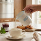 White ceramic milk jug with blue rim pouring milk into a teacup at breakfast