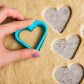 Hand holding a blue heart shape cookie cutter above rolled dough with cut heart cookies