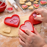 Hand pressing a red heart shaped cookie cutter into dough with stamped heart cookies on a cooling rack
