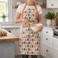 Woman wearing an orange and brown leaf print kitchen apron with front pocket while whisking batter in a bowl in a bright home kitchen