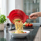 Red silicone strainer with a steel handle pouring cooked spaghetti into a white bowl.
