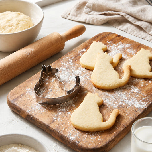 Stainless steel cat cookie cutter with cut cat cookies and a rolling pin on a wooden board