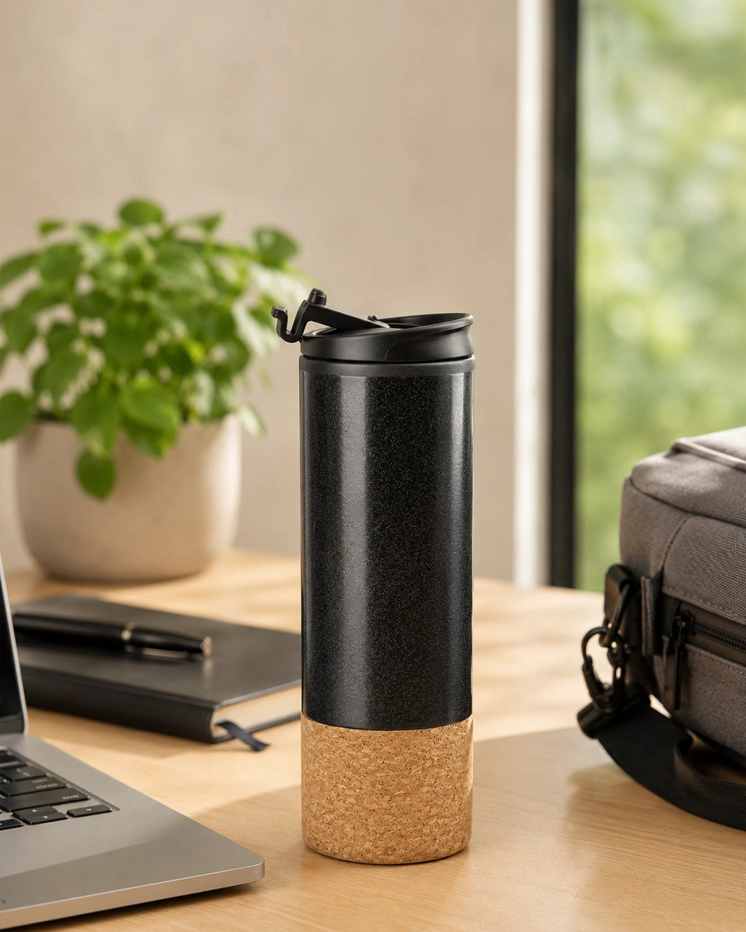 Black travel mug with cork base on a desk beside a laptop and work bag