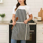 white and black apron with front pocket worn by woman holding wooden spoon, striped cotton kitchen apron for women