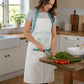 Woman wearing a white cotton kitchen apron with teal Country Kitchen farmhouse print and teal pocket trim while chopping vegetables on a wooden board in a country style kitchen